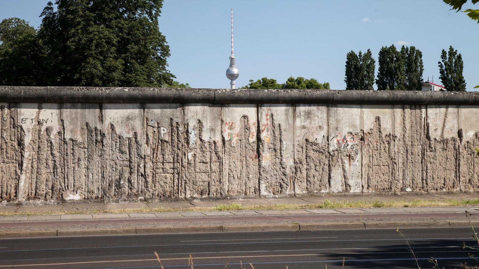 Die Mauer in der Bernauer Straße. Der Putz ist größtenteils abgeschlagen, sodass die Stahlarmierung sichtbar ist. Im Hintergrund ist der Fernsehturm erkennbar.